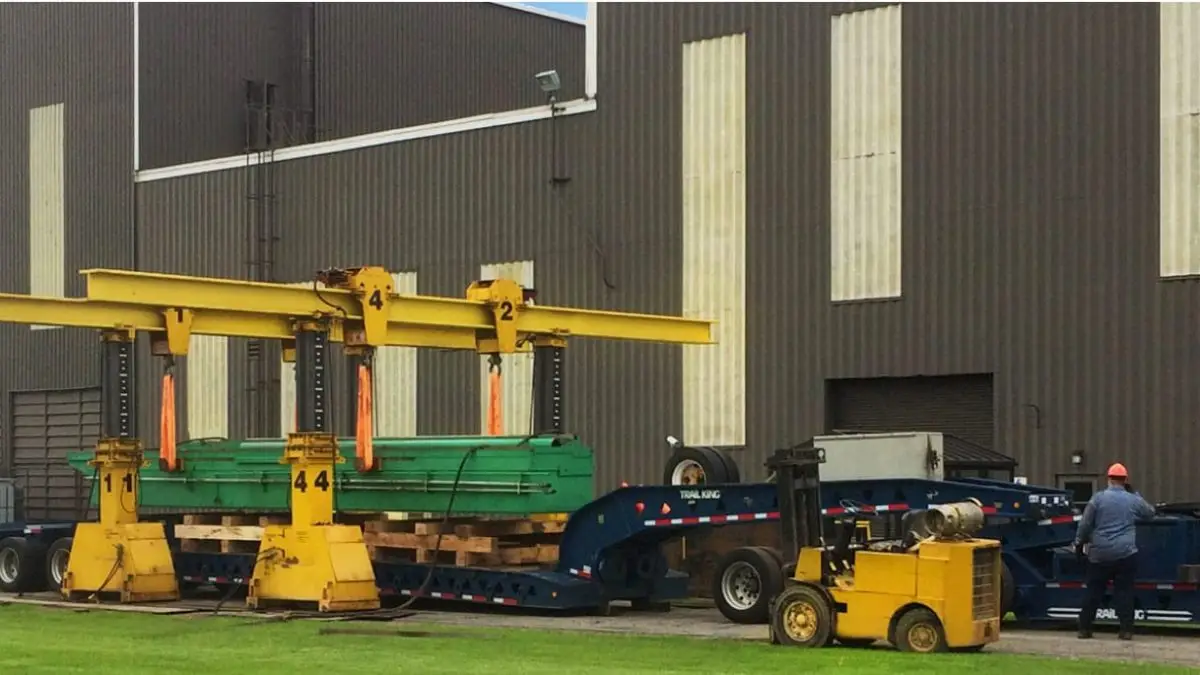 A large industrial machine is being lifted onto a flatbed trailer by two yellow gantry cranes outside a factory building, with workers and equipment nearby on a grassy area. - JV Rigging A large industrial machine is being lifted onto a flatbed trailer by two yellow gantry cranes outside a factory building, with workers and equipment nearby on a grassy area.