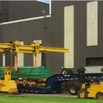 A large industrial machine is being lifted onto a flatbed trailer by two yellow gantry cranes outside a factory building, with workers and equipment nearby on a grassy area. - JV Rigging A large industrial machine is being lifted onto a flatbed trailer by two yellow gantry cranes outside a factory building, with workers and equipment nearby on a grassy area.