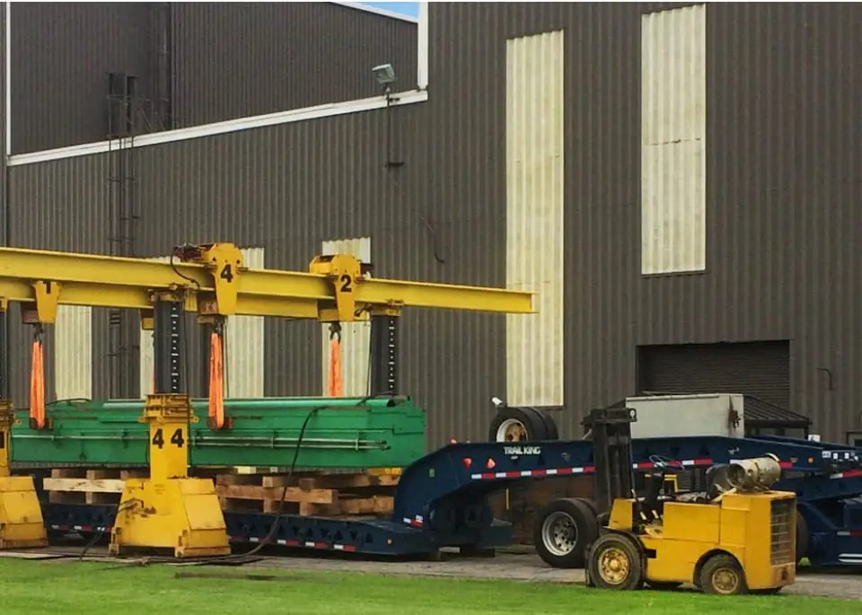 A large industrial machine is being lifted onto a flatbed trailer by two yellow gantry cranes outside a factory building, with workers and equipment nearby on a grassy area.